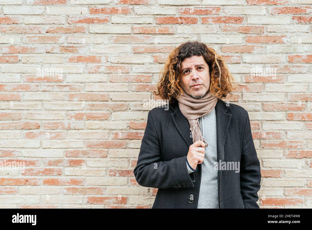 Male in smart casual clothes with curly hair touching scarf and looking at camera while standing against brick wall on street Stock Photo