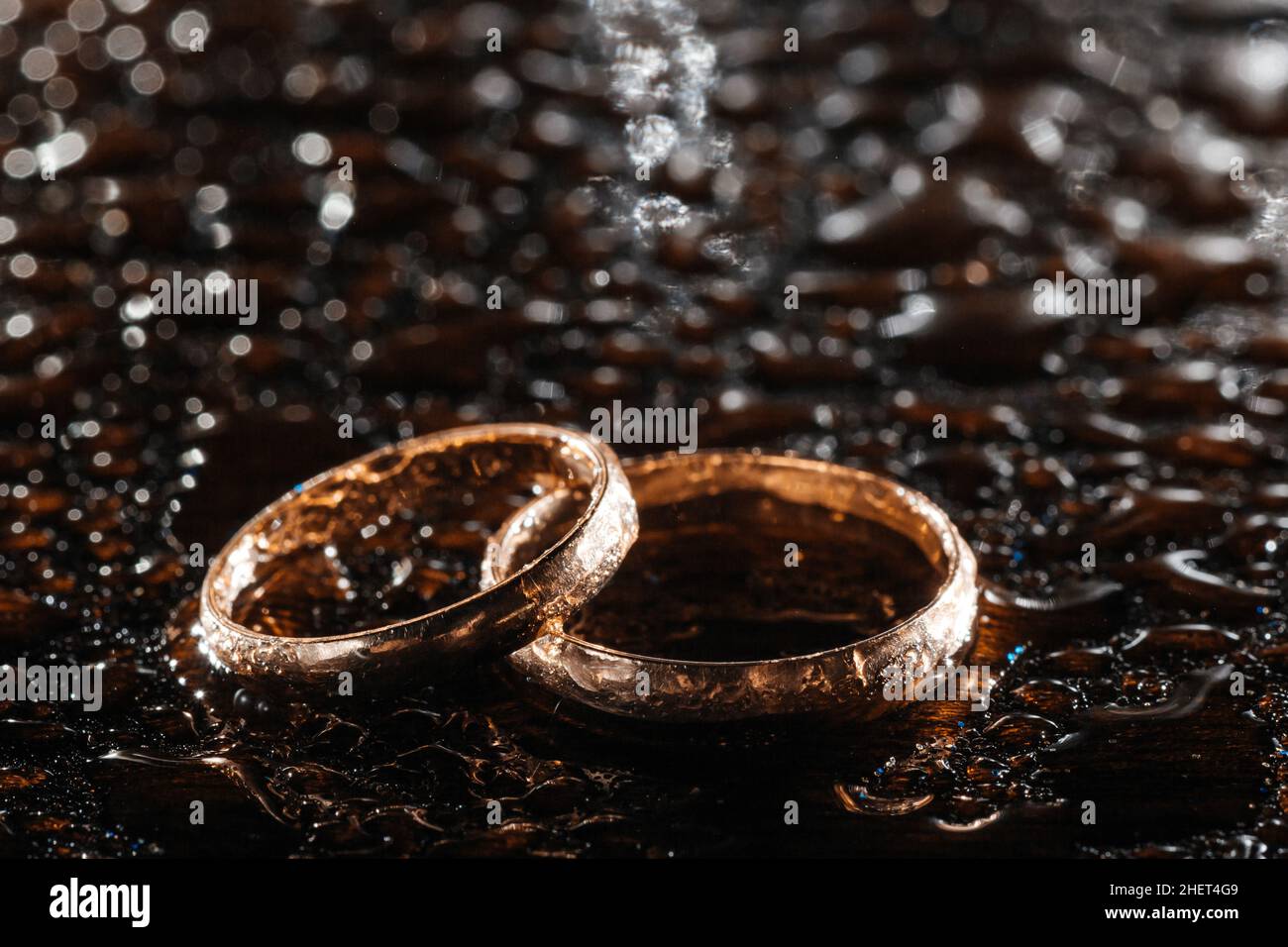 Two gold wedding rings in rain drops on a black wet background. Stock Photo