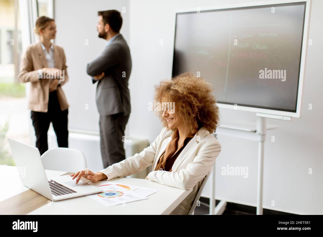 Young curly hair businesswoman using laptop in the office with young people works behind her Stock Photo