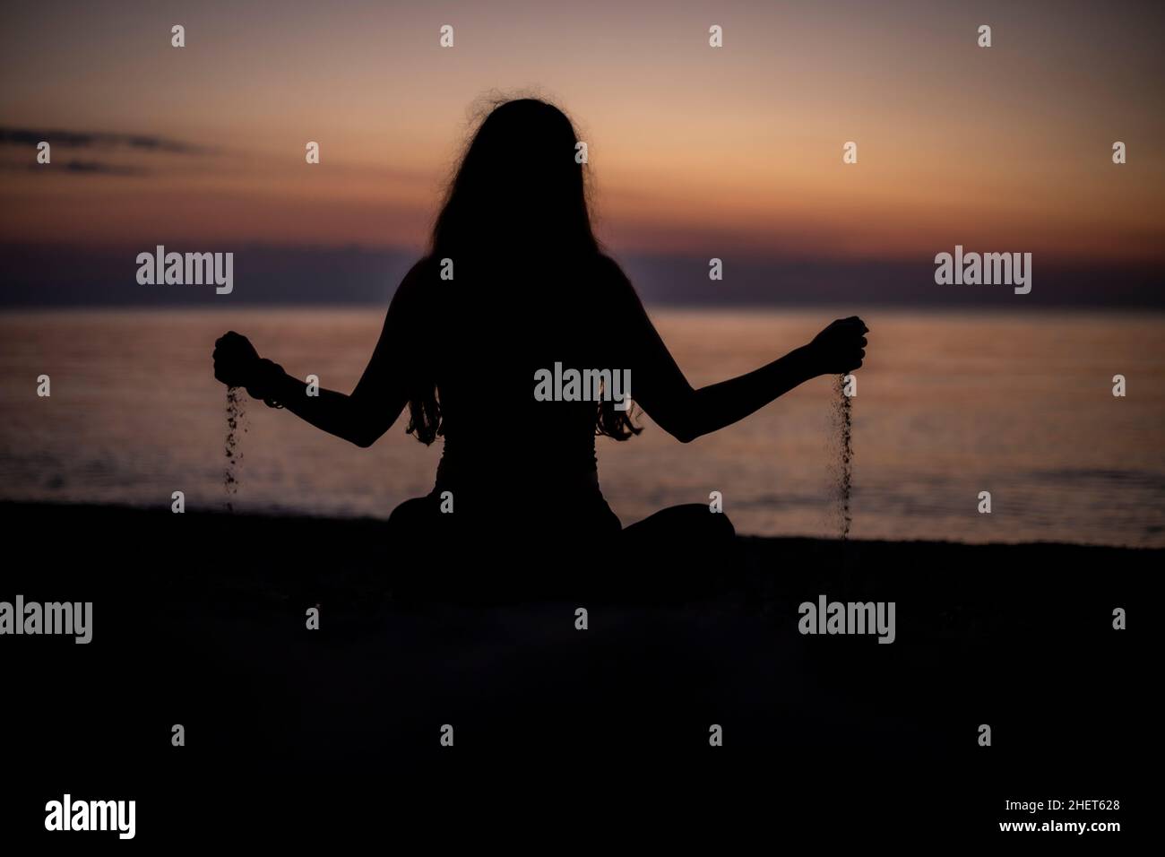 Rear view of a silhouette of a woman playing with sand sitting on a beach at sunset Stock Photo