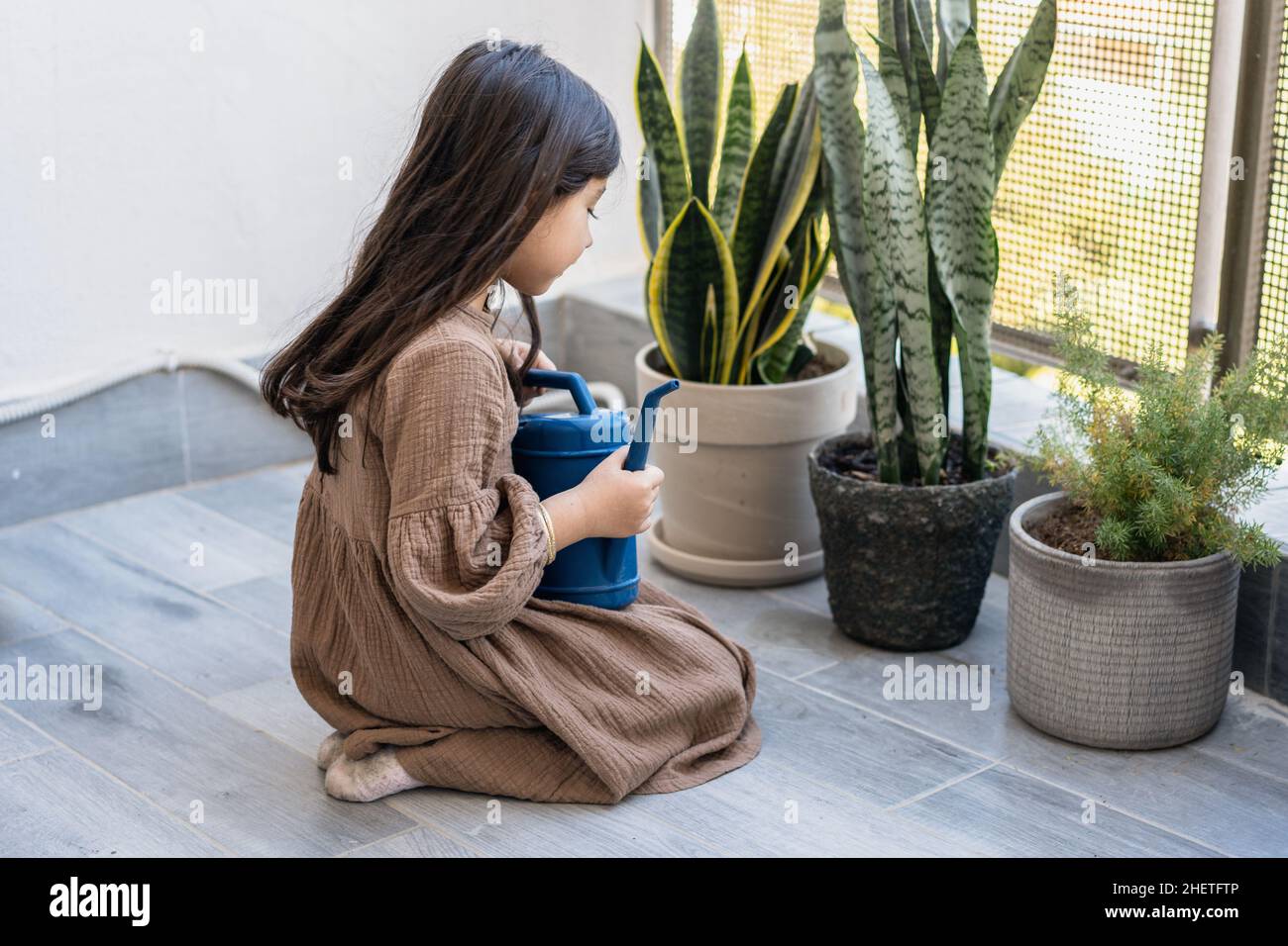 Little girl is watering flowers on the balcony at home. Active preschool child watering plants with water can. Stock Photo