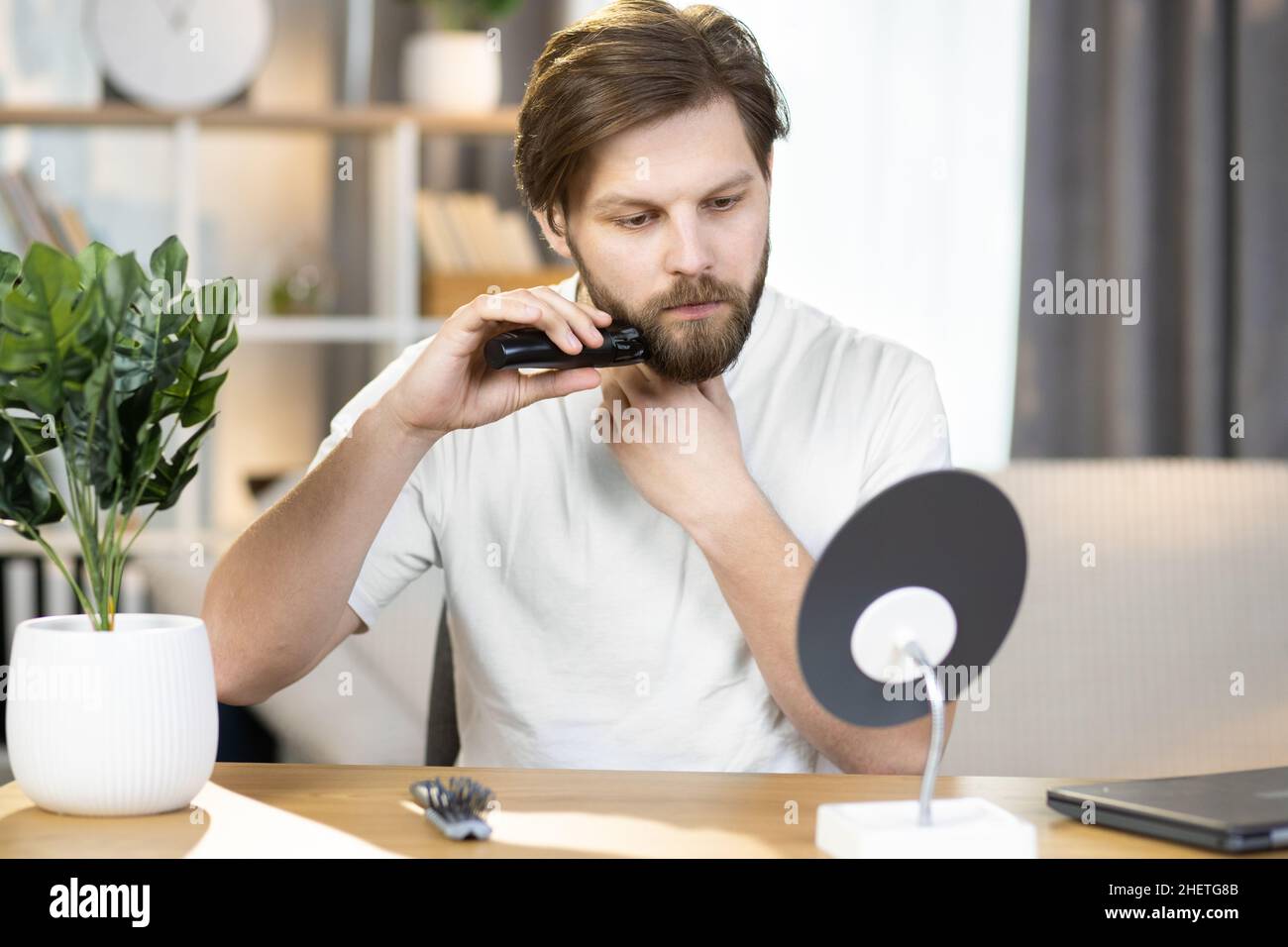 Young handsome caucasian man performs hygienic procedures at home. The bearded guy tidies up his beard while shaving it with an electric razor Stock Photo