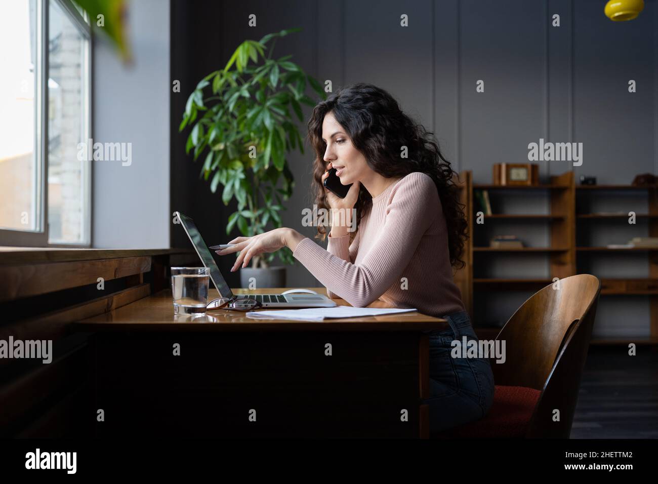 Young female entrepreneur talking on mobile phone and pointing at laptop screen, working in office Stock Photo