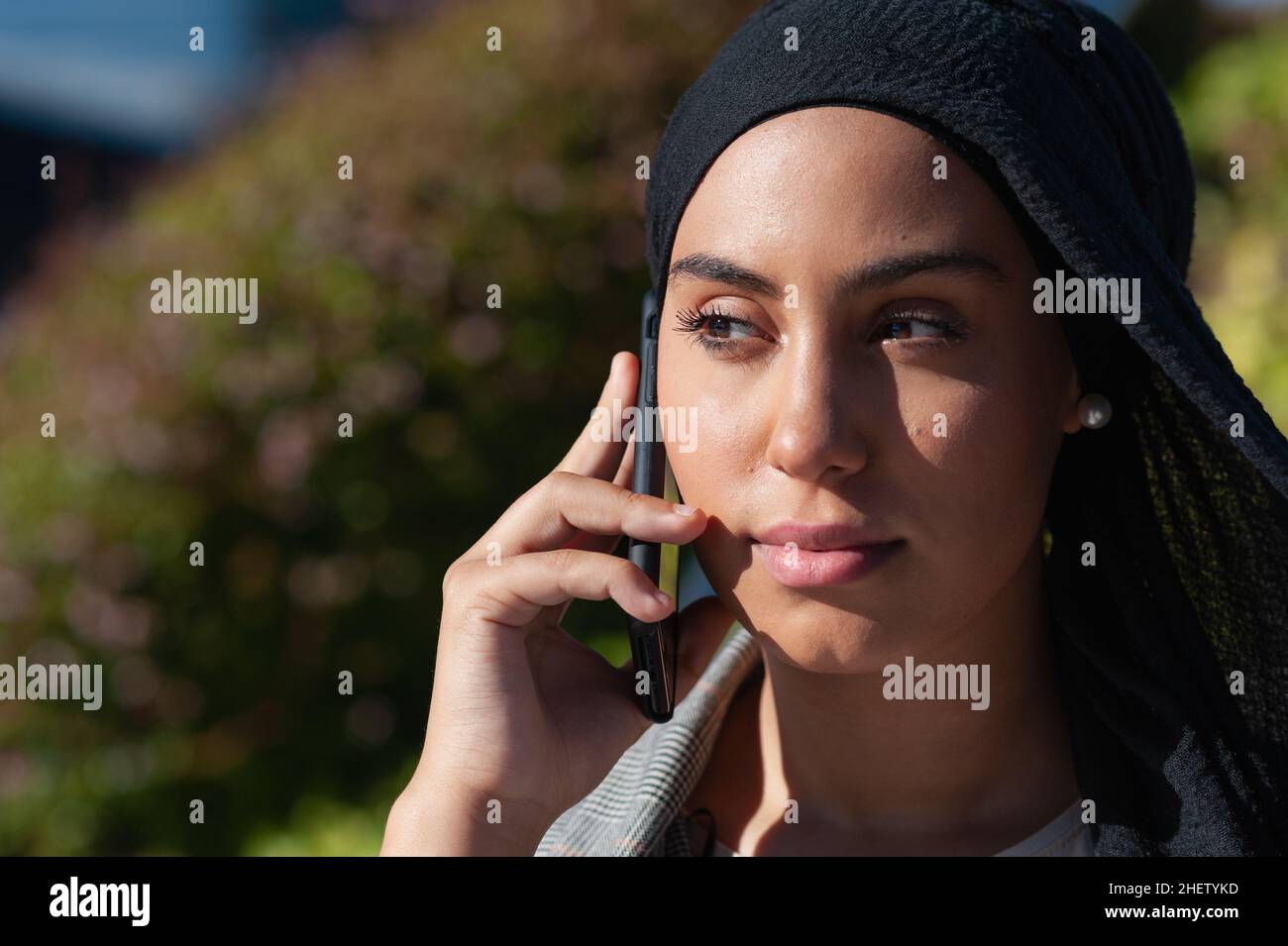 Close up view of an unsmiling muslim woman talking with her phone while looking at camera in the park in a sunny day. Stock Photo
