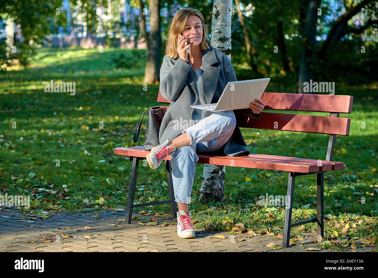 A woman with a laptop is sitting on a park bench on a sunny summer day Stock Photo