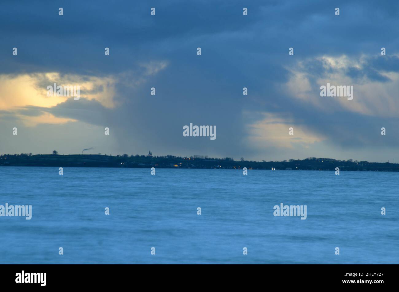 Evening sky with a large cloud formation that shows precipitation stripes, the lighs of a settlement can be seen at the horizon Stock Photo