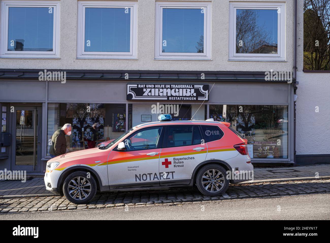 A parked German Red Cross emergency ambulance with doctor is parked in front of a funeral home business. An elderly man, wearing a Covid mask passes. Stock Photo