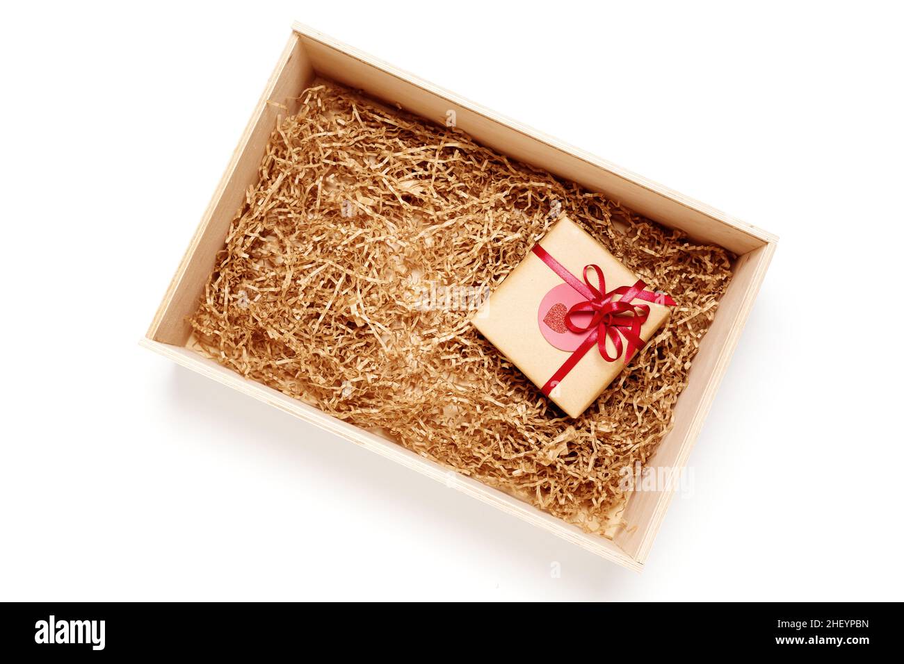 Wooden box with present wrapped in brown craft paper with red ribbon, filled with paper filler, top view from above on white background Stock Photo