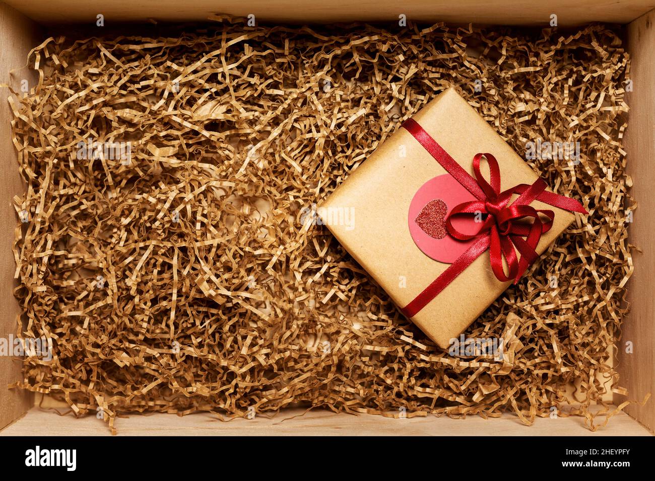 Wooden box with present wrapped in brown craft paper with red ribbon, filled with paper filler, top view from above Stock Photo