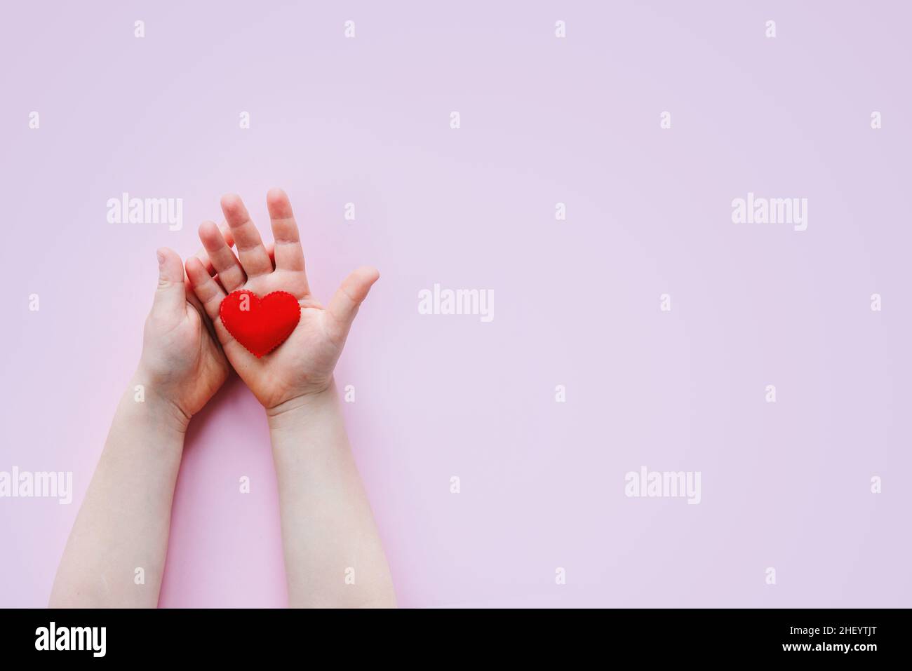 Childs hand holding a red fabric heart on pink background with copy ...