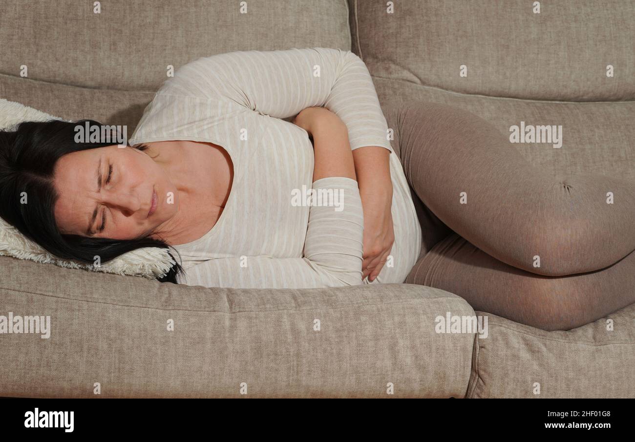 Middle aged mature woman suffering from stomach ache at home. Abdominal pain, food poisoning, diarrhea, stomach ulcer, menstruation pain, menopause. Stock Photo