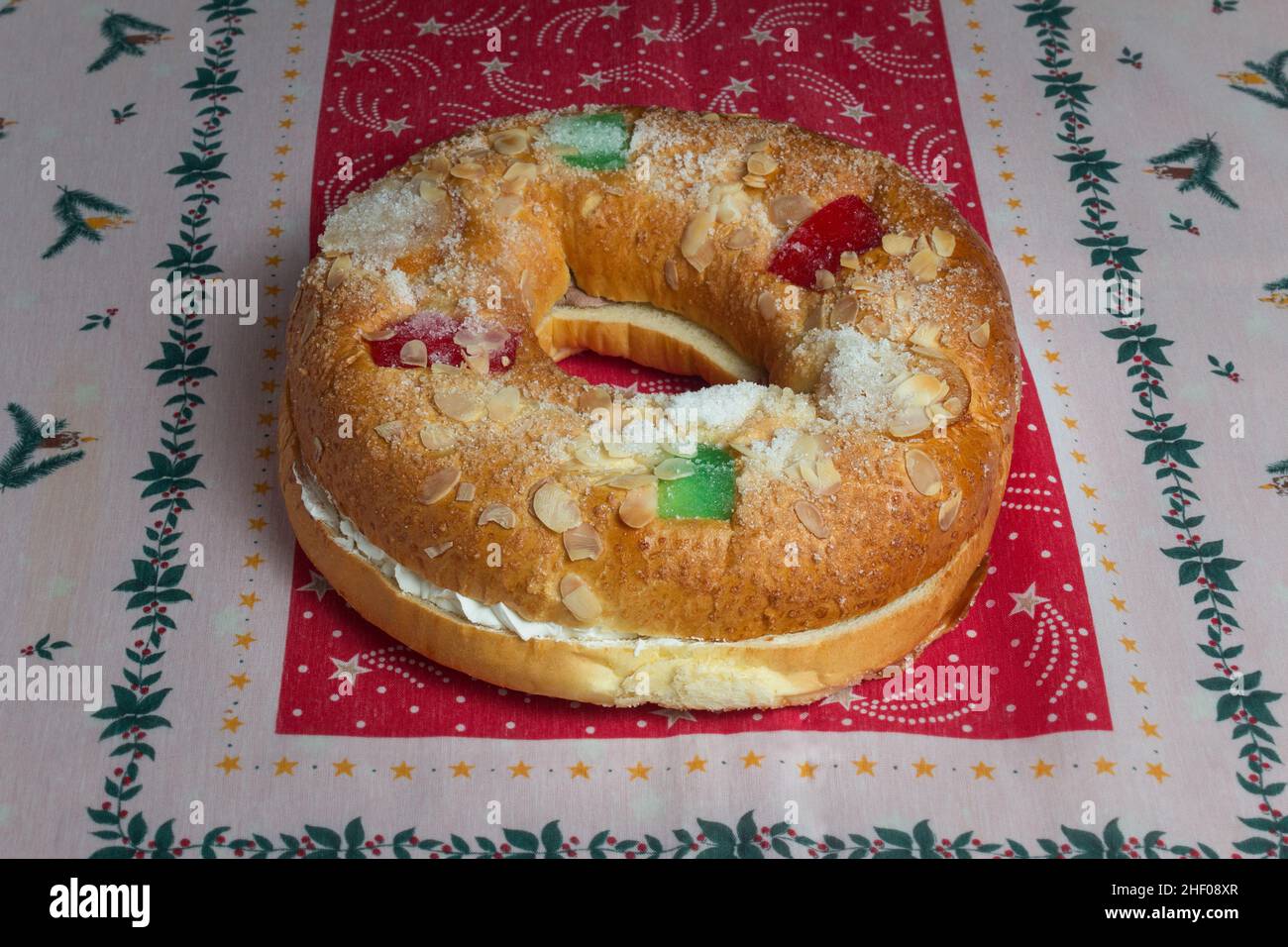 An oval-shaped cake with a sweet filling with sweet whipped cream decorated with almonds and candied fruit on a tablecloth with Christmas decoration. Stock Photo