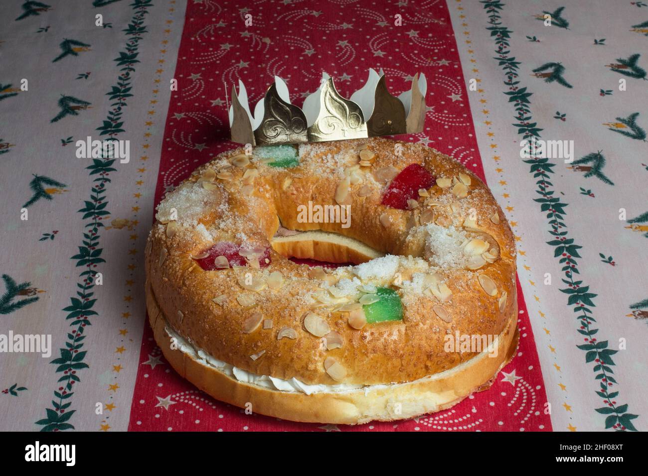 A large donut cake with a sweet creamy filling inside, almonds, candied fruit adorning the top with a crown of kings on a tablecloth decorated with Ch Stock Photo