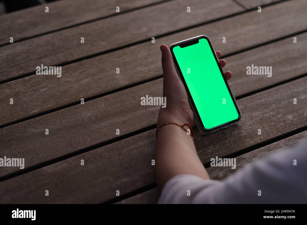 over the shoulder view of hand holding green screen mobile phone on wooden cafe table at night Stock Photo