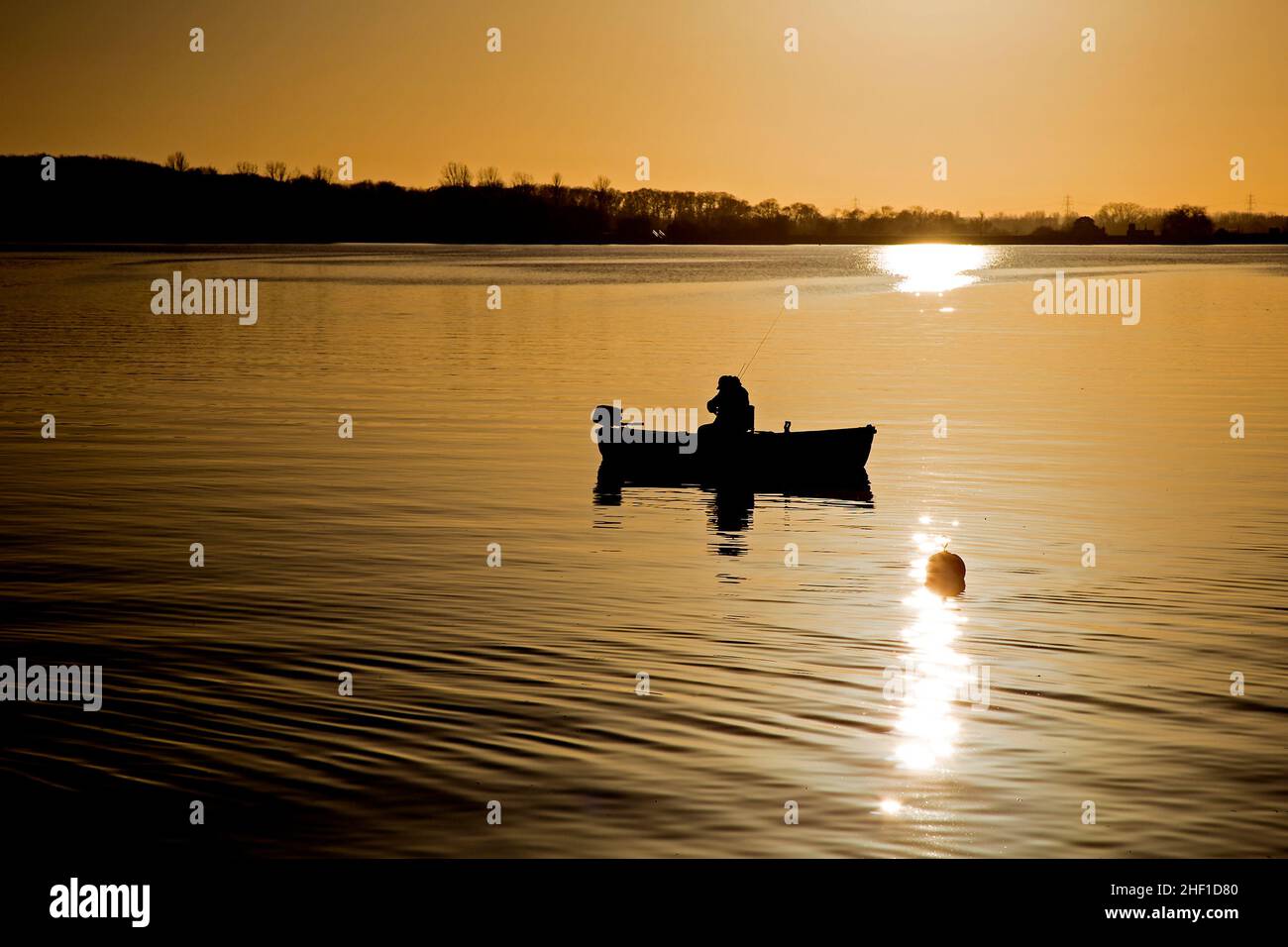 A silhouette of a fisherman in a small boat on a reservoir at sunset ...