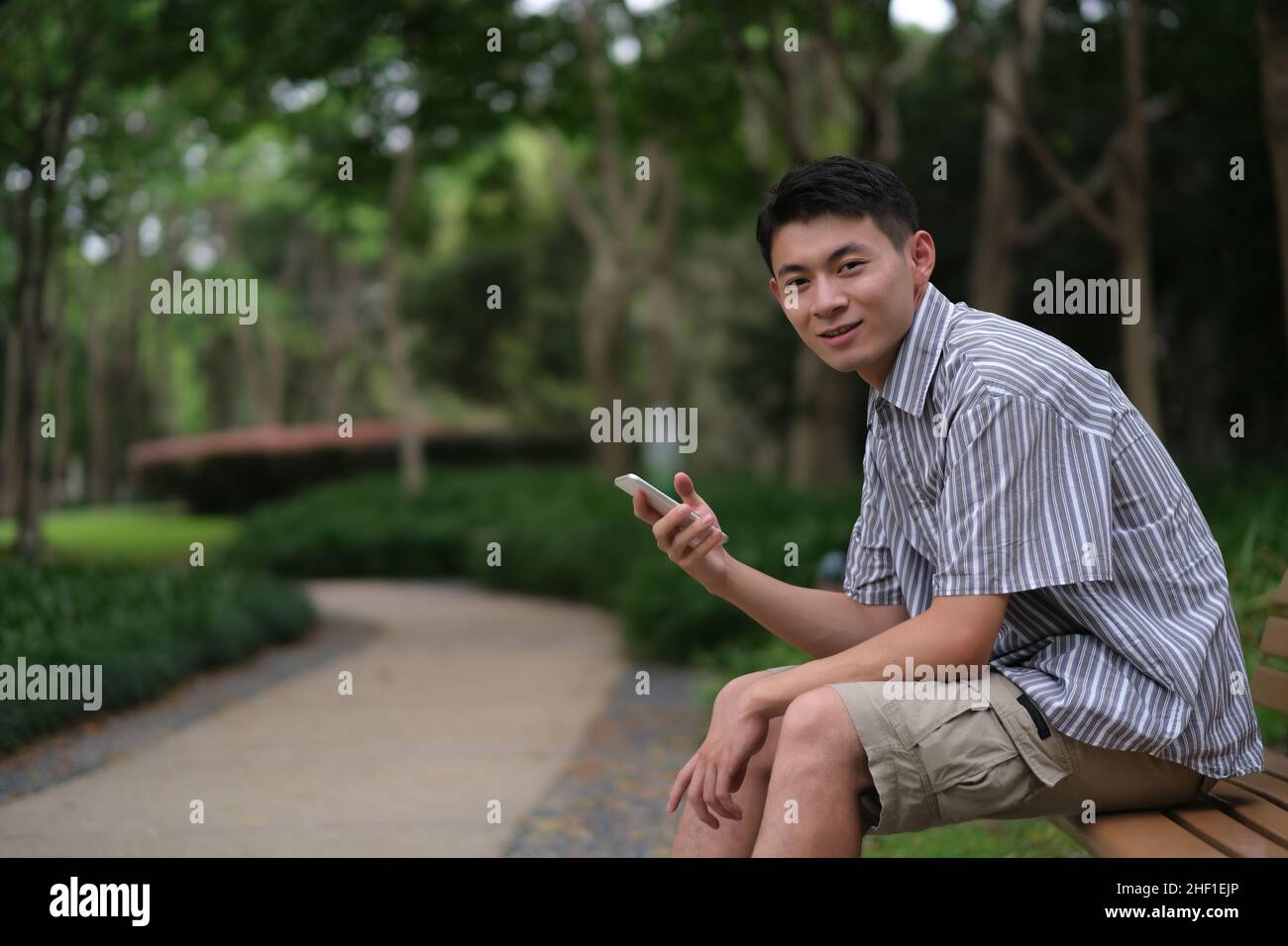 one Asian young man smiling and using phone, looking at camera in natural green park Stock Photo