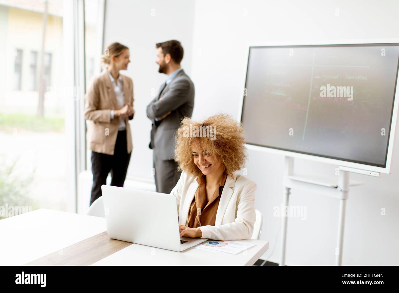 Young curly hair businesswoman using laptop in the office with young people works behind her Stock Photo