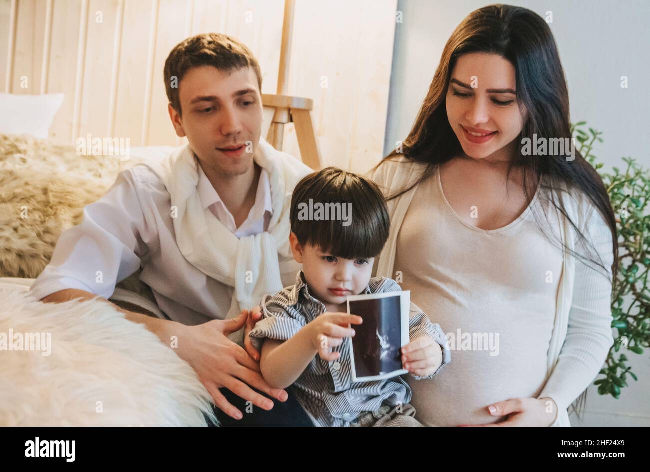 Happy family with child expecting for new baby. Pregnant mother, father and little kid son with ultrasound image in hands sitting together at home. Mo Stock Photo