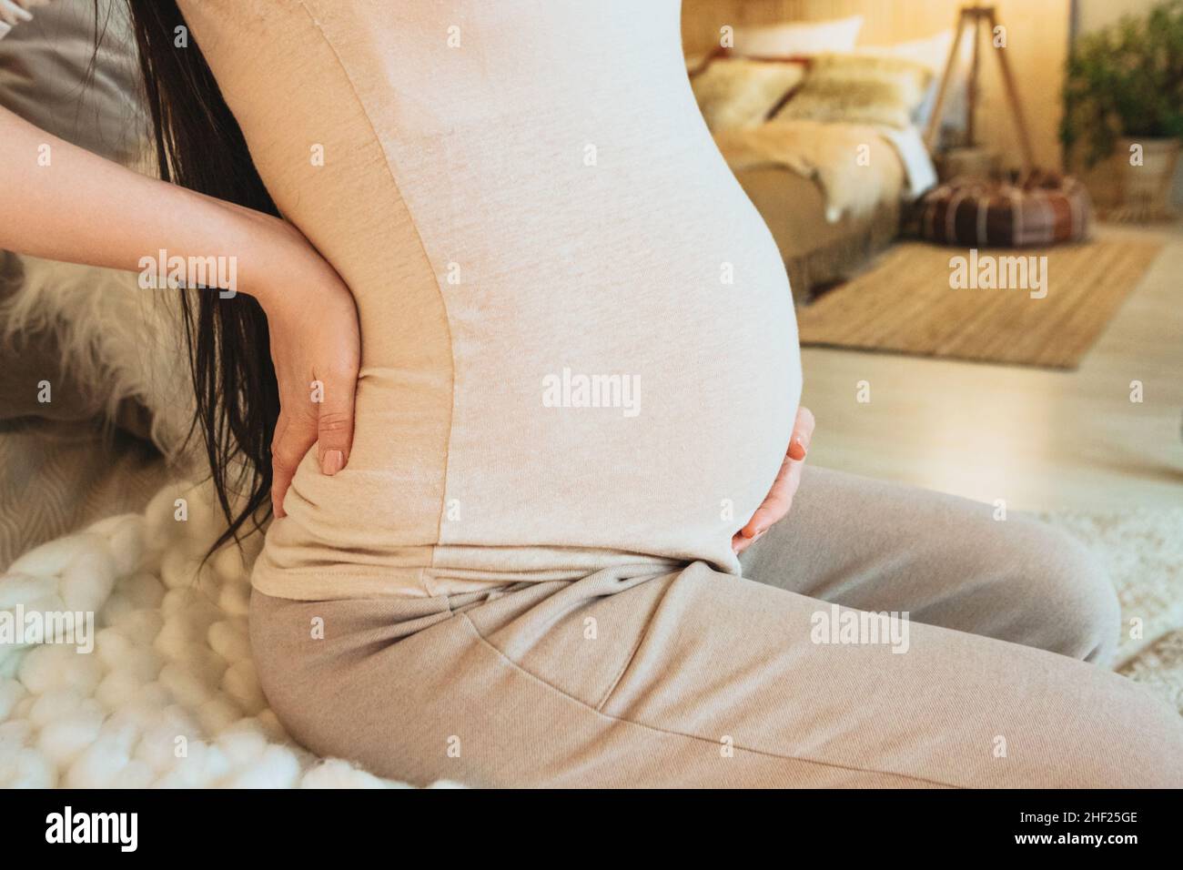 Close up photo of pregnant woman holding belly while sitting on couch at home, woman expecting baby gently touching tummy, cropped. Pregnancy, materni Stock Photo