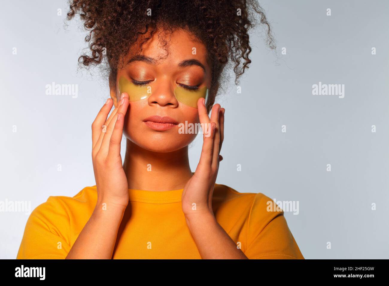 Skincare concept. Young calm african american woman enjoying beauty routine, applying hydrogel cosmetic eye patches to under-eye area on face, isolate Stock Photo