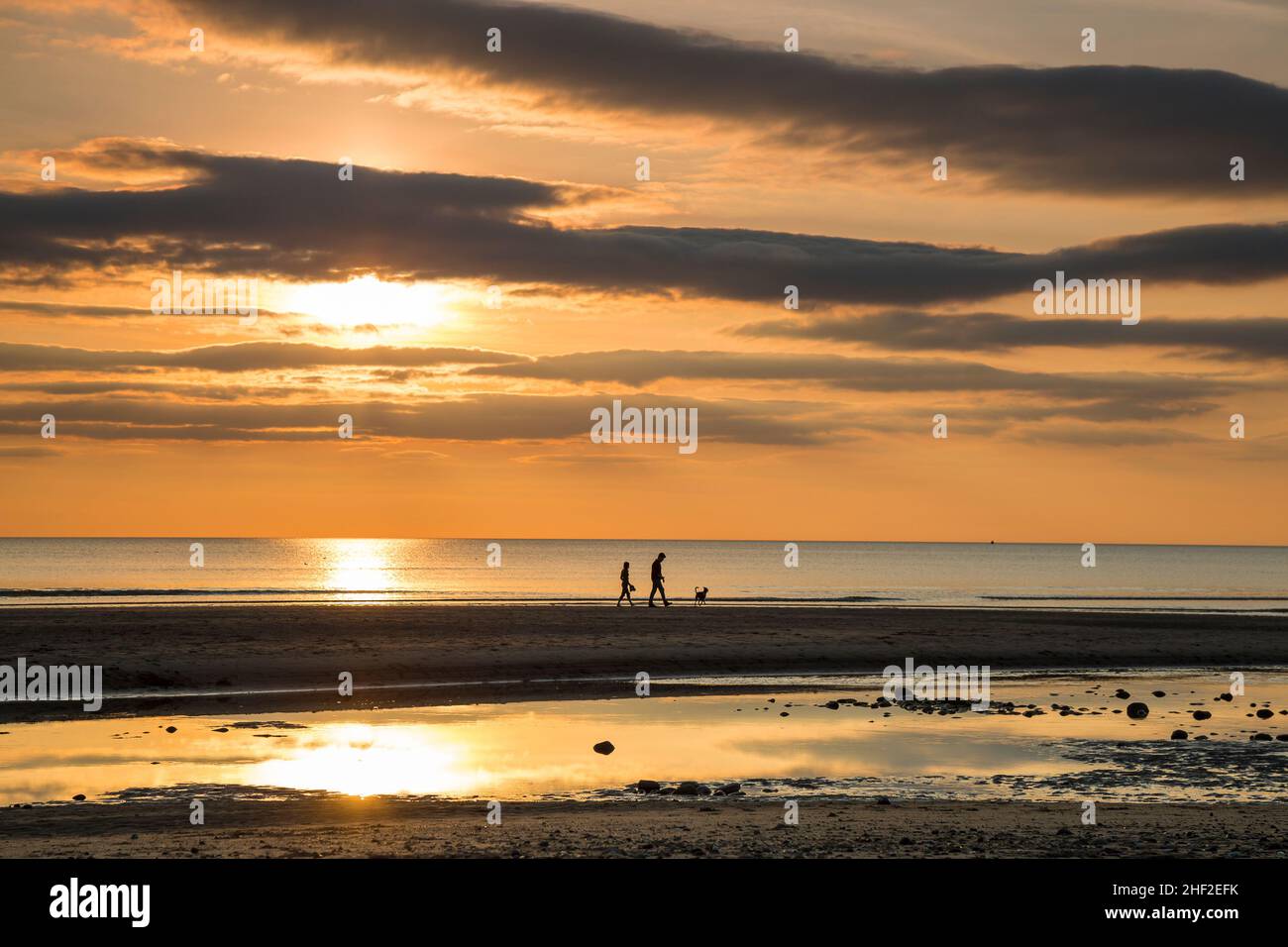 UK sunset beach landscape with young couple and pet dog silhouetted in the distance, walking on the shoreline sand. Stock Photo