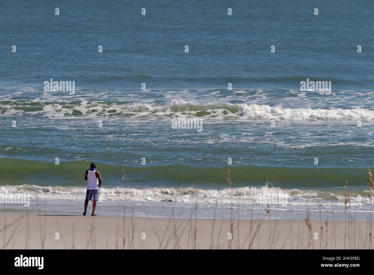 A black man stands alone on a beach looking out at the vast and restless sea Stock Photo