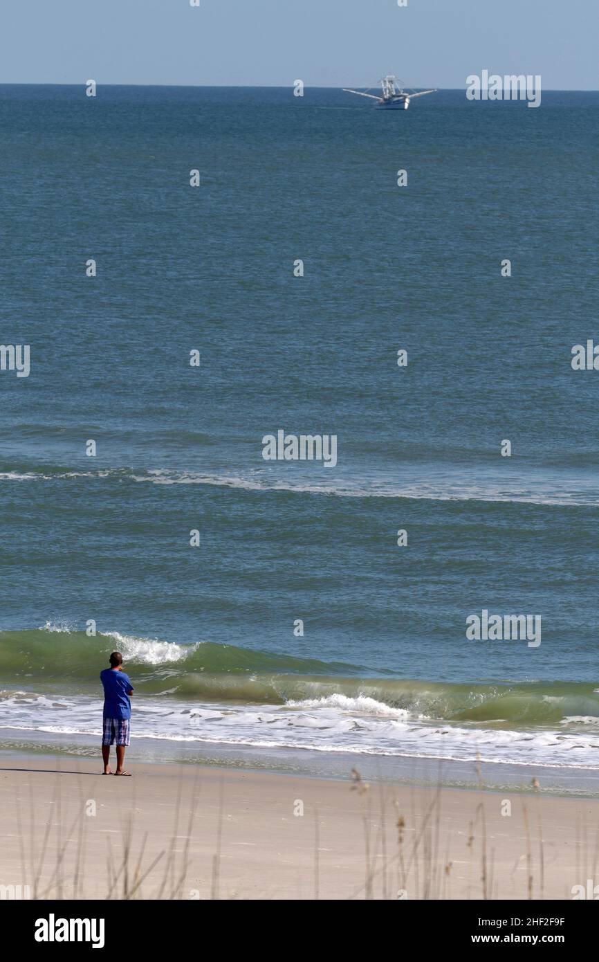 A thoughtful black man stands on the beach looking out to sea as a Trawler moves past in the distance Stock Photo