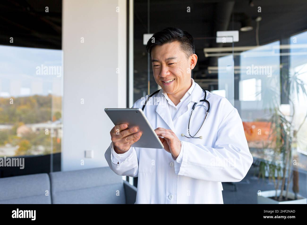 Cheerful and friendly Asian in a modern clinic, online video call, uses a tablet, to communicate with patients, and online meetings with colleagues Stock Photo