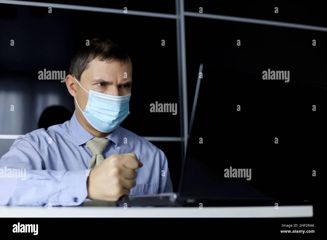 Angry man in face mask and office clothes banging his fist on the table while sitting at laptop. Concept of disgruntled boss during online meeting Stock Photo