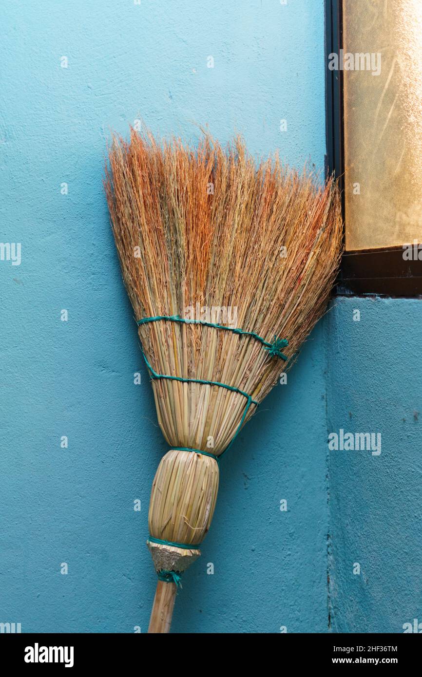 A hand made straw broom leans against a turquoise interior wall beside a black window in Jinotega, Nicaragua Stock Photo