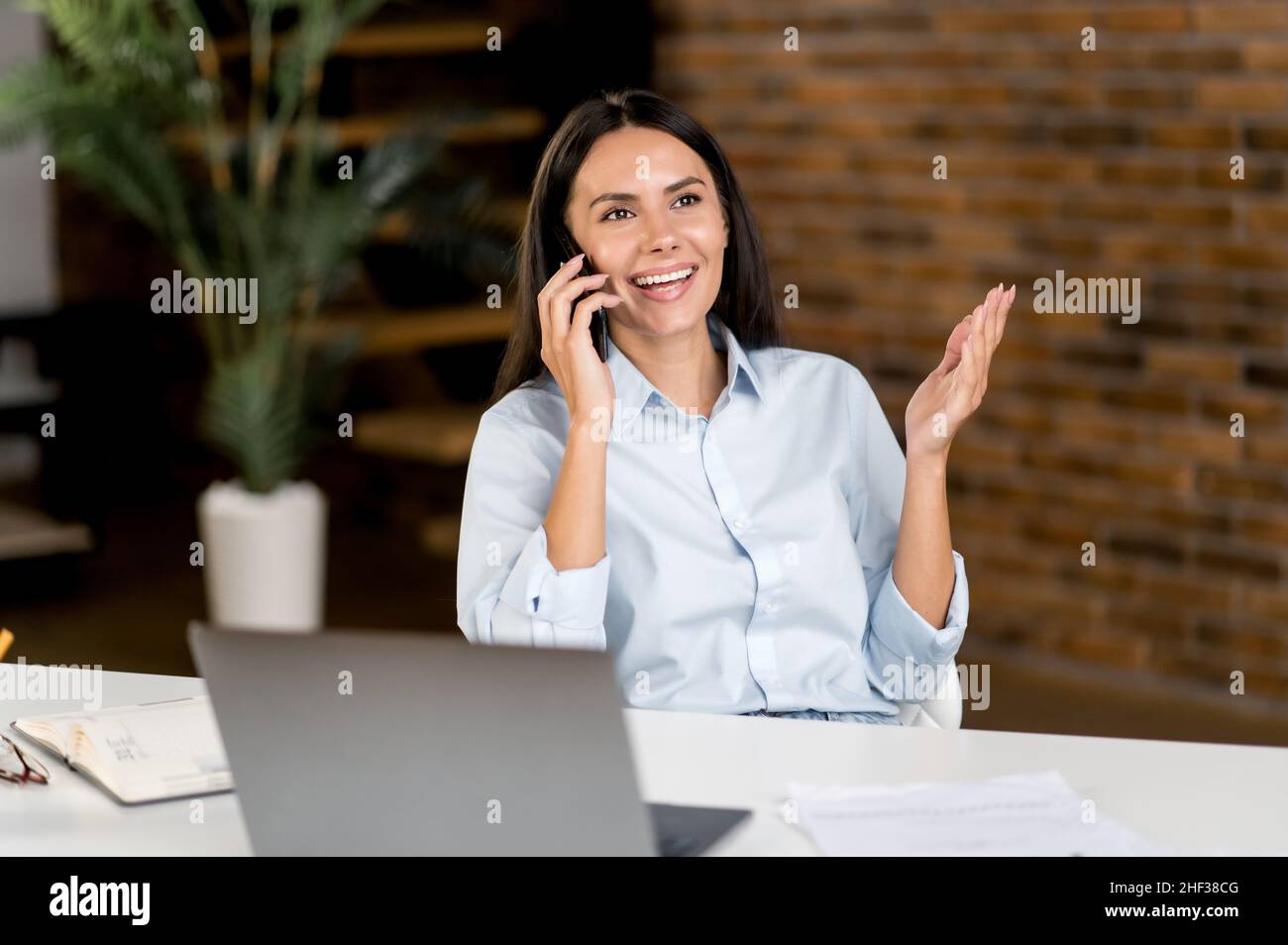 Smart successful positive Caucasian brunette, IT specialist, sales manager, sits at work desk, talking on smartphone with friend or client while working, smiling friendly, gesturing with his hands Stock Photo