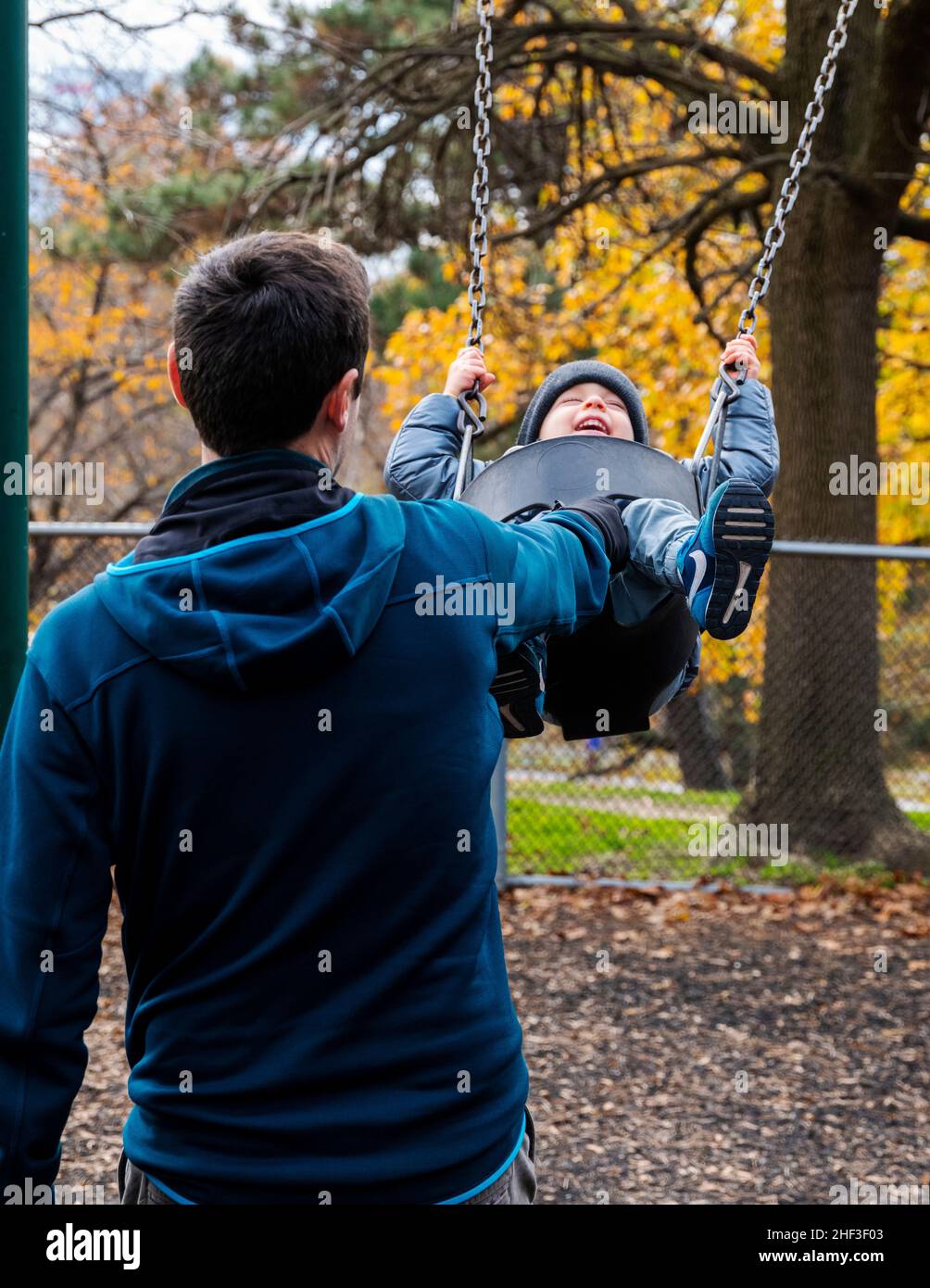 Father pushing two year old boy swinging on city park swing set; Corinthian Gardens