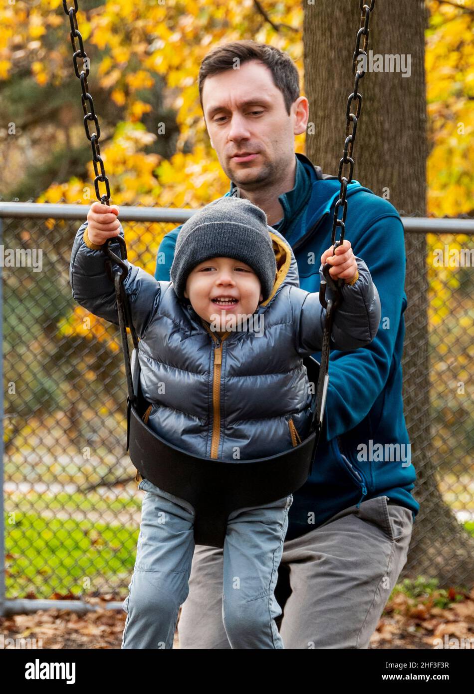 Father pushing two year old boy swinging on city park swing set; Corinthian Gardens; Philadelphia; Pennsylvania; USA Stock Photo