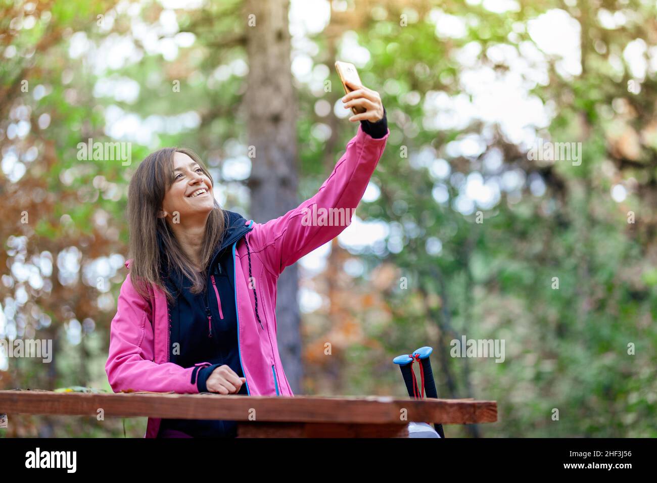 Hiker girl resting on a bench in the forest. Backpacker with pink jacket taking selfie with smartphone. Stock Photo
