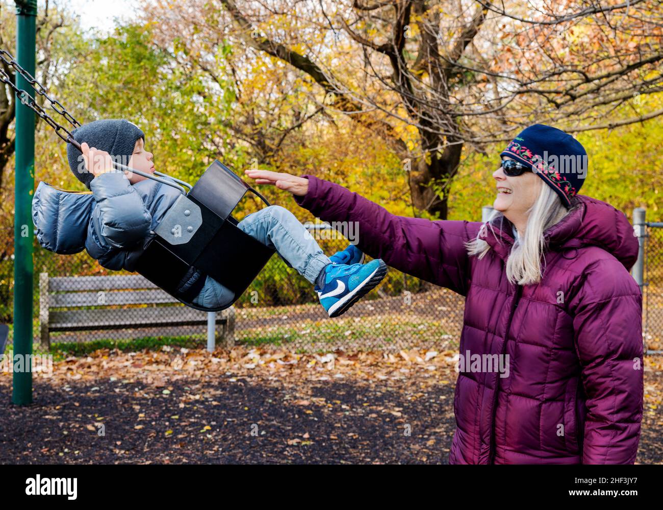 Senior grandmother pushing two year old boy swinging on city park swing set; Corinthian Gardens; Philadelphia; Pennsylvania; USA Stock Photo