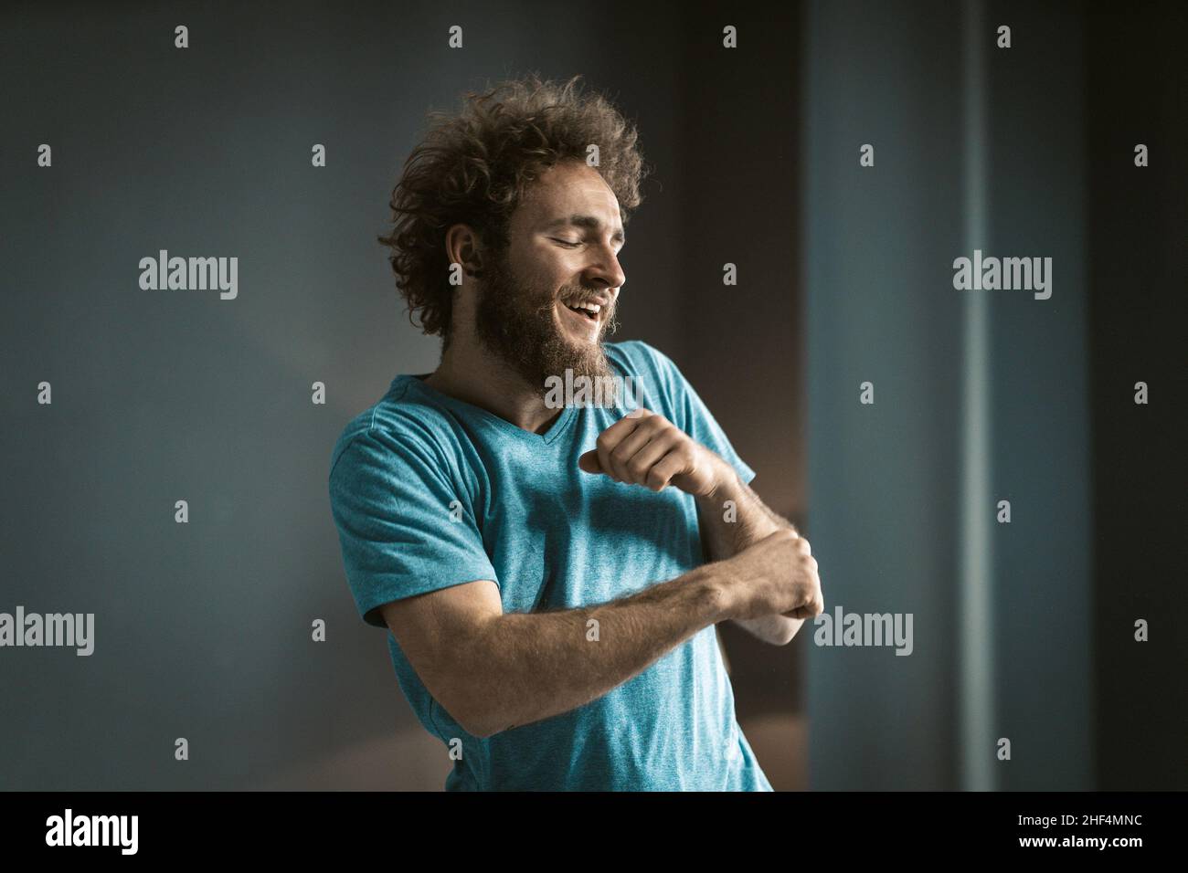 Coily Guy in a Blue T-shirt and a Tattoo on his Neck Makes Funny Movements by his Hands. Young Man Dancing in the Room at Home.Copy Space. Close-up. High quality photo Stock Photo
