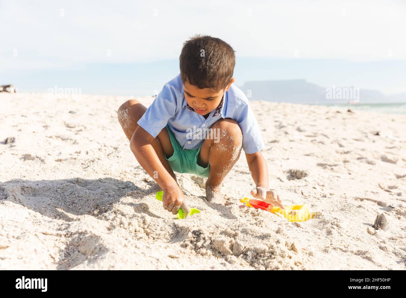 Full length of biracial boy crouching while playing with sand and toys at beach on sunny day Stock Photo