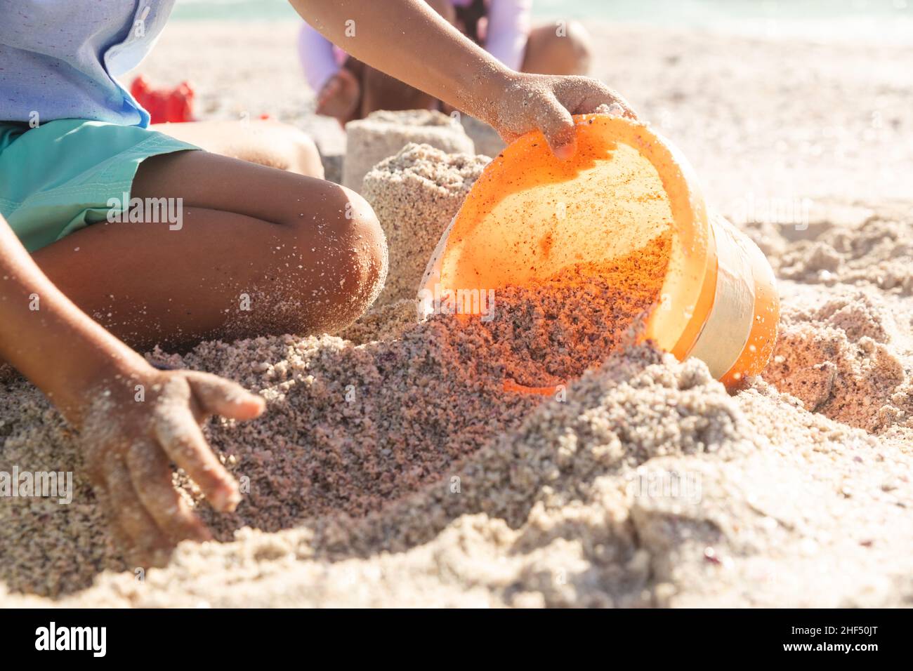 Midsection of biracial boy collecting sand in bucket with sister playing at beach on sunny day Stock Photo