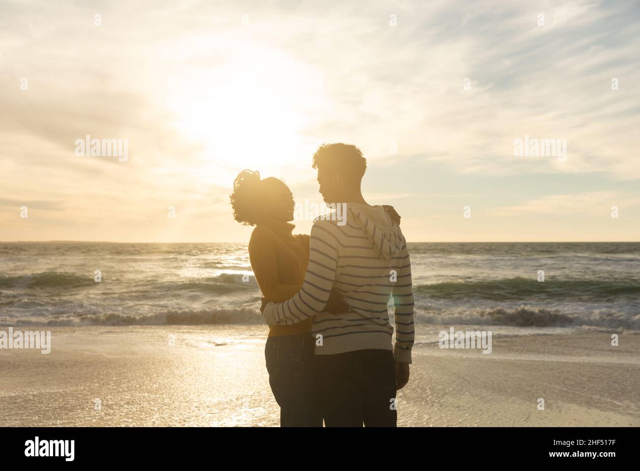 Romantic biracial couple embracing while standing at beach against sky during sunset Stock Photo
