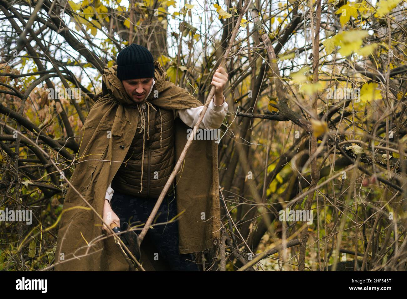 Portrait of serious tourist male wearing raincoat tent chopping small branches with small shovel in forest on overcast cold day. Stock Photo