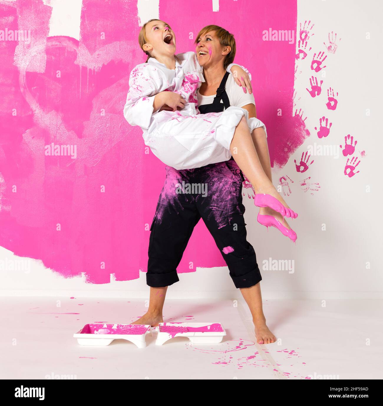 Mother and daughter standing in front of a wall painted with pink color.Mother is holding the girl and and the girl has color on both feet. Both are w Stock Photo