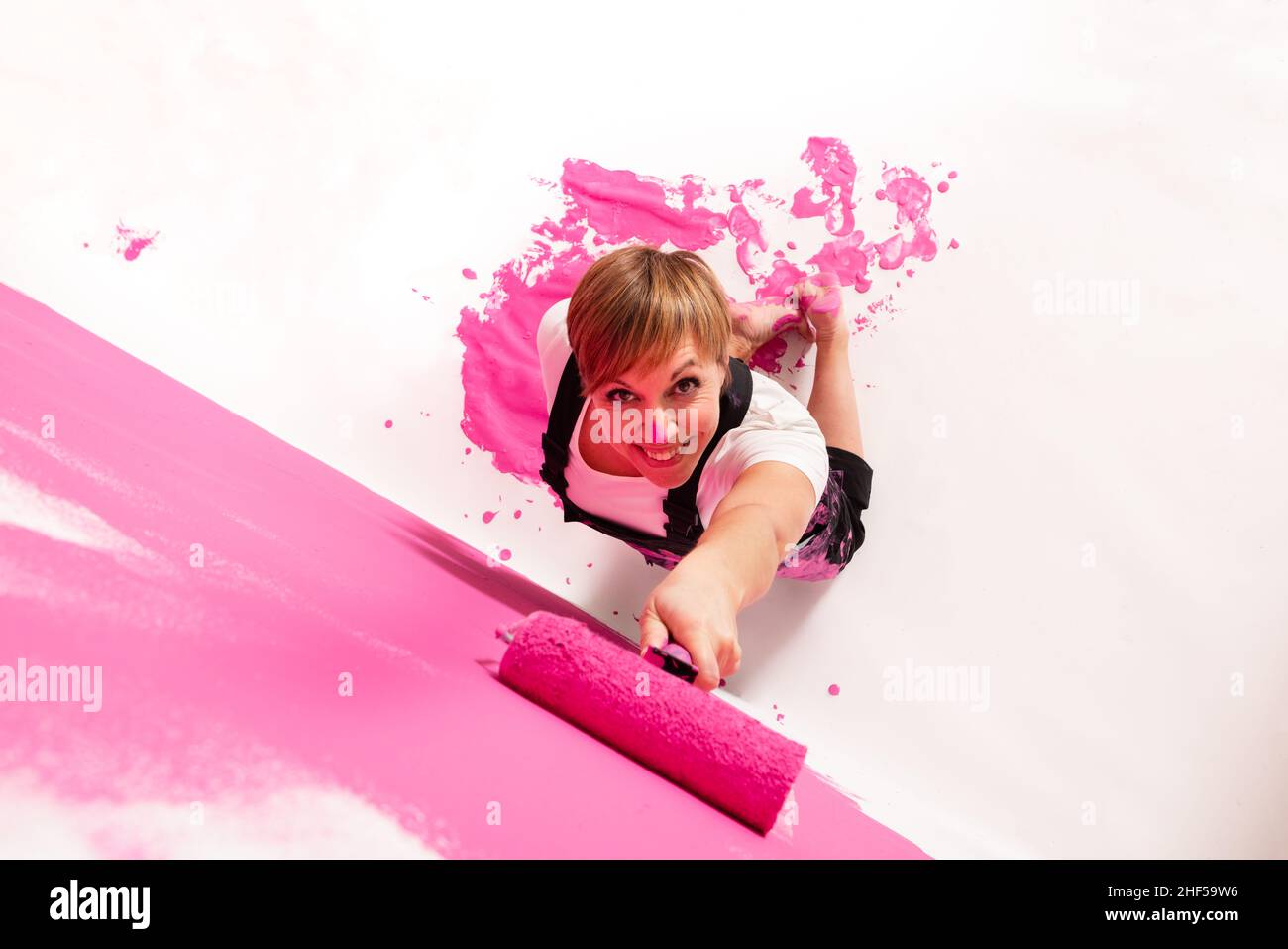 Young woman painting a wall with pink colored roller. She is seen from above and she is looking to the camera with a friendly smile. Stock Photo
