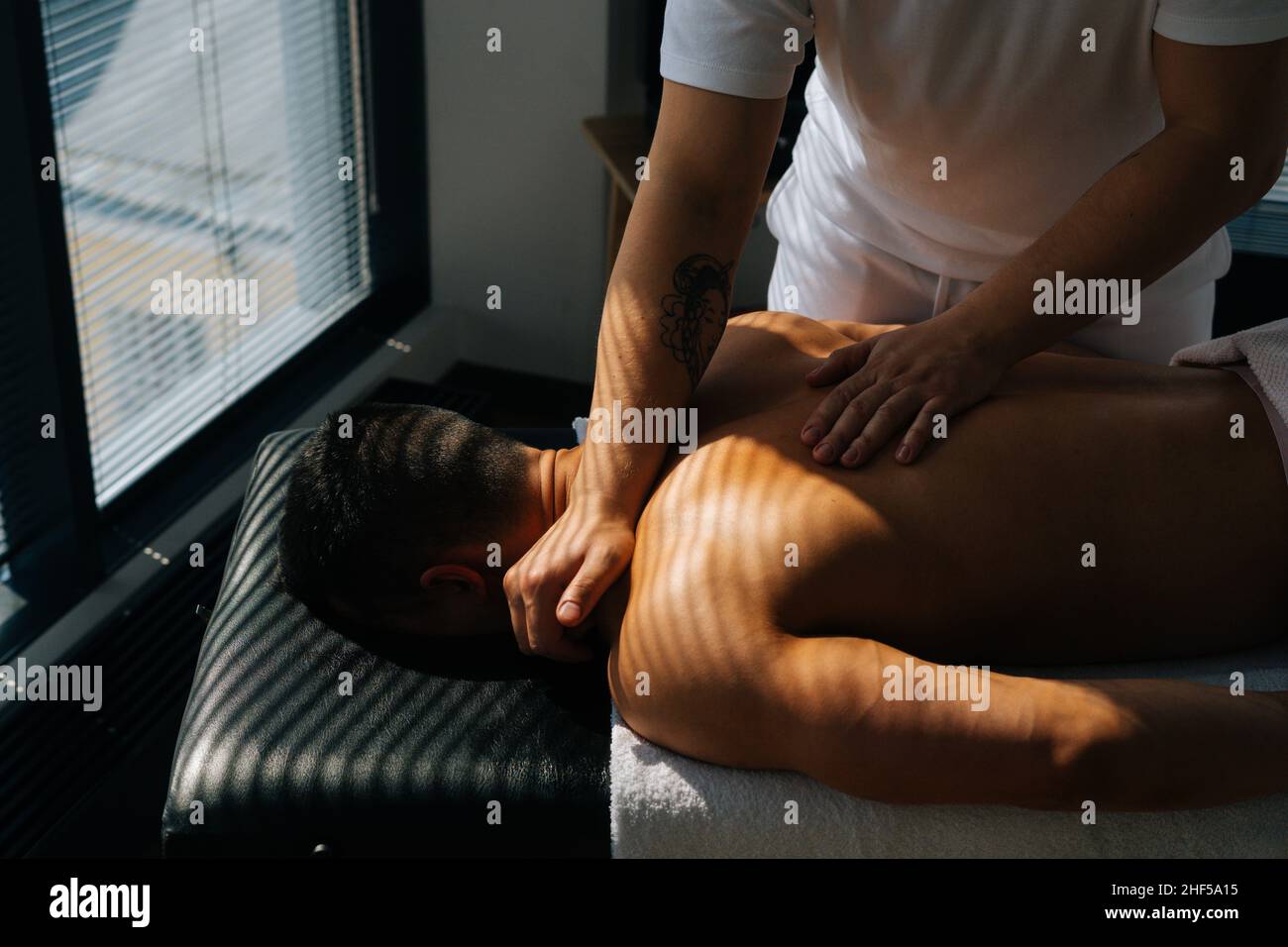 Close-up top view of professional male masseur with strong tattooed hands massaging neck and shoulders of muscular sports man lying on stomach Stock Photo