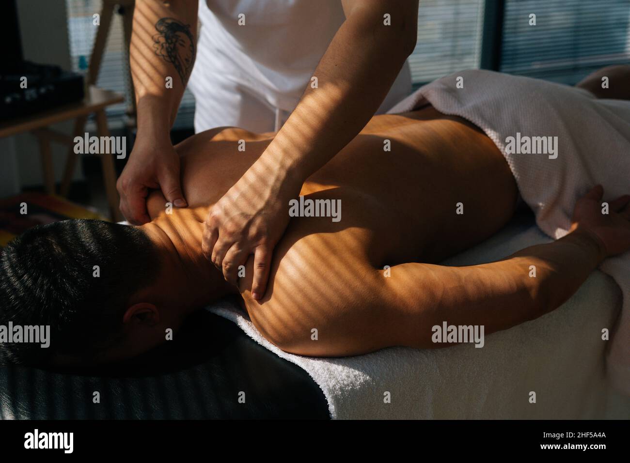 Top close-up view of unrecognizable male masseur with strong tattooed hands massaging neck and shoulders of muscular sports man lying on stomach, Stock Photo