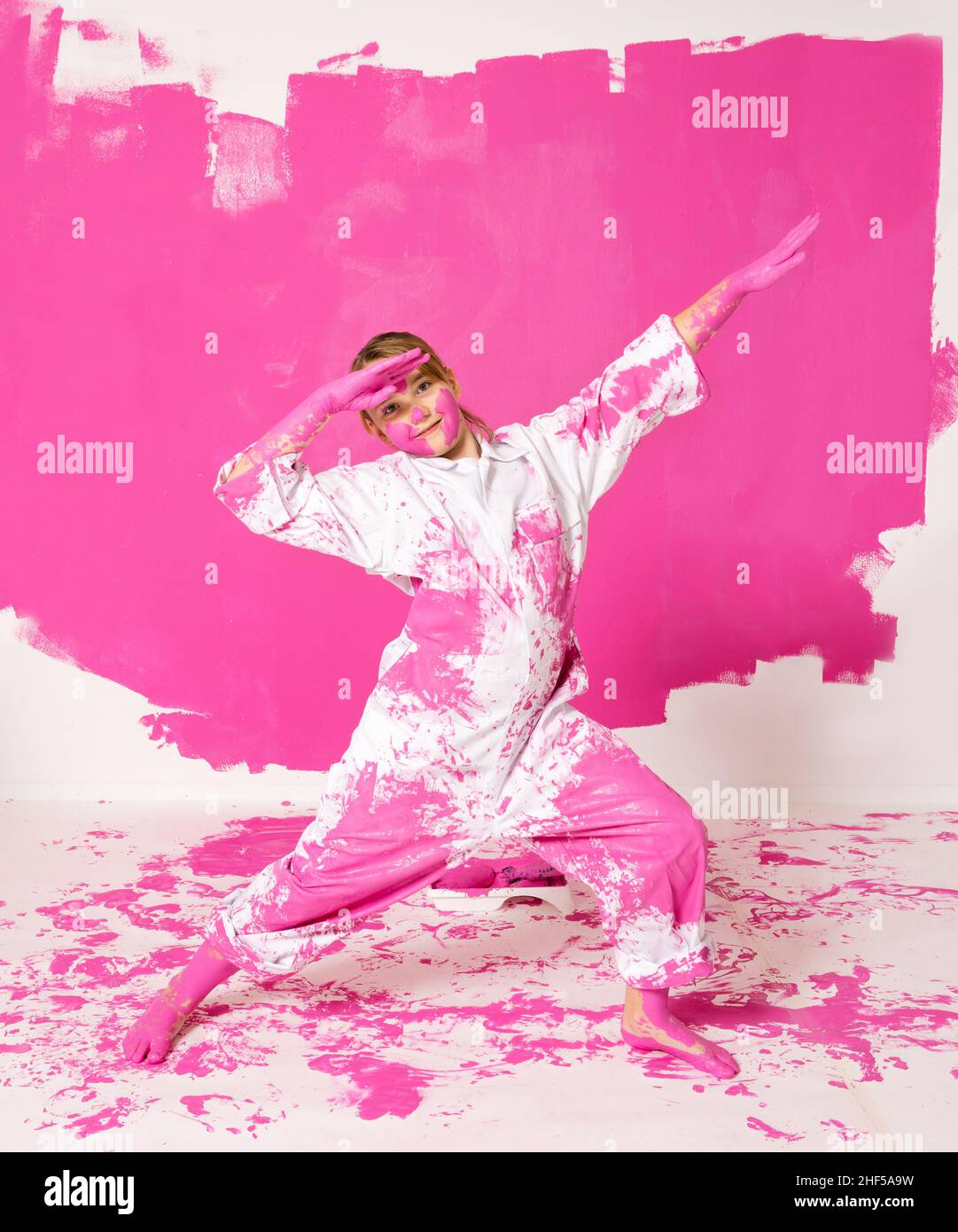 Young girl having fun painting a wall with pink color. Traces of hands and feet are seen on the wall behond her. She is wearing working clothes and he Stock Photo
