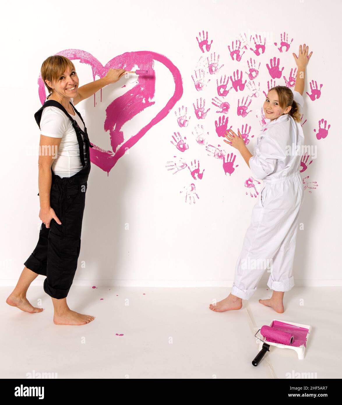 Mother and 10 year old daughter is painting with pink color on white wall. They are both looking into camera with happy smiles. Stock Photo