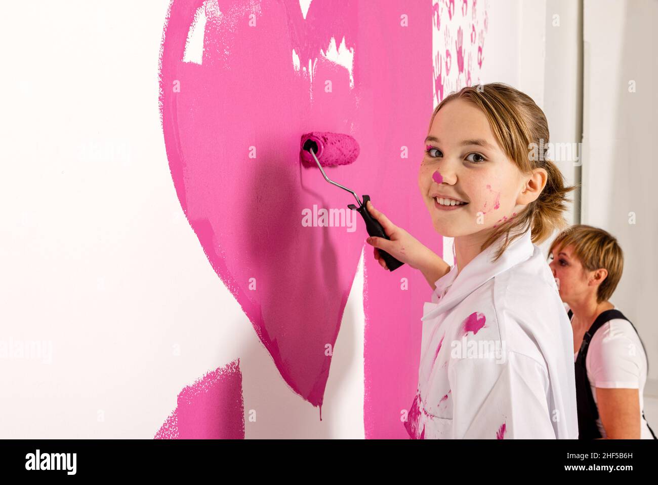 Young girl is painting with a roller filled with pink paint on a wall. Girl is looking at the camera with a happy smile. Mother is seen in the backgro Stock Photo