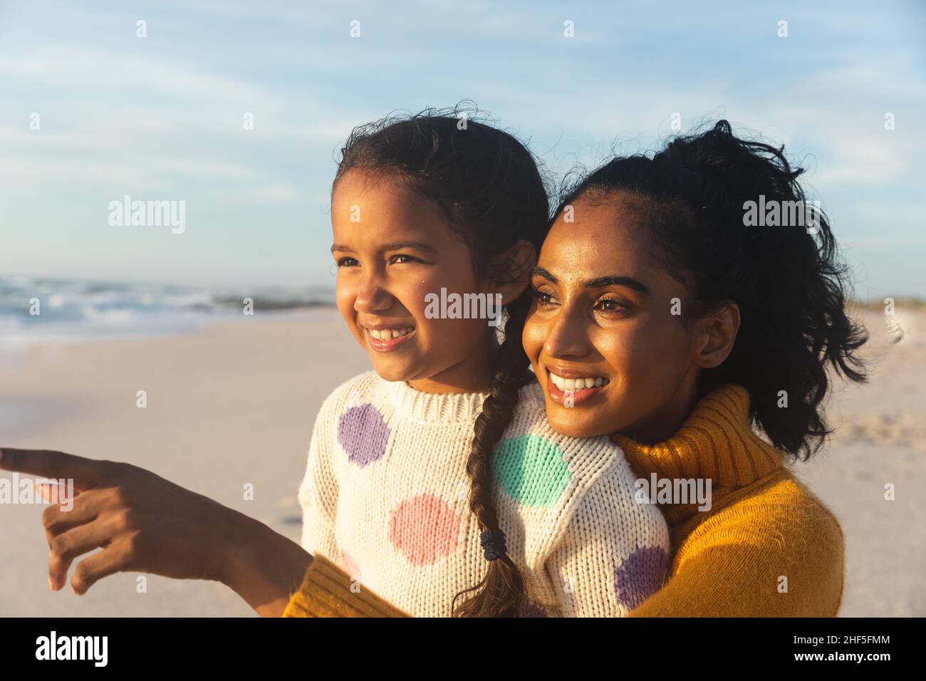 Happy biracial woman pointing to daughter at beach against sky during sunset Stock Photo