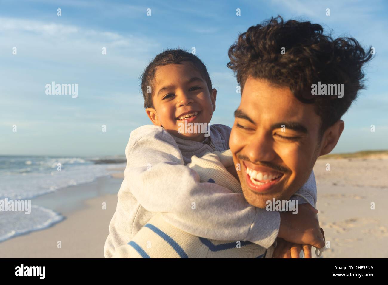 Close-up of happy biracial father giving son piggyback ride to son at beach against sky Stock Photo