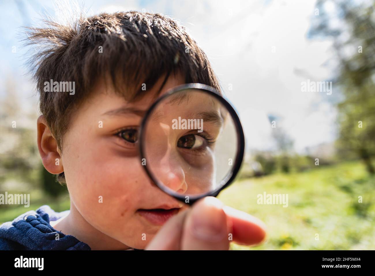 Curious child exploring with a magnifying glass with focus on his eye. Kid 's face magnified by a magnifying lens conveys curiosity, discovery concept Stock Photo